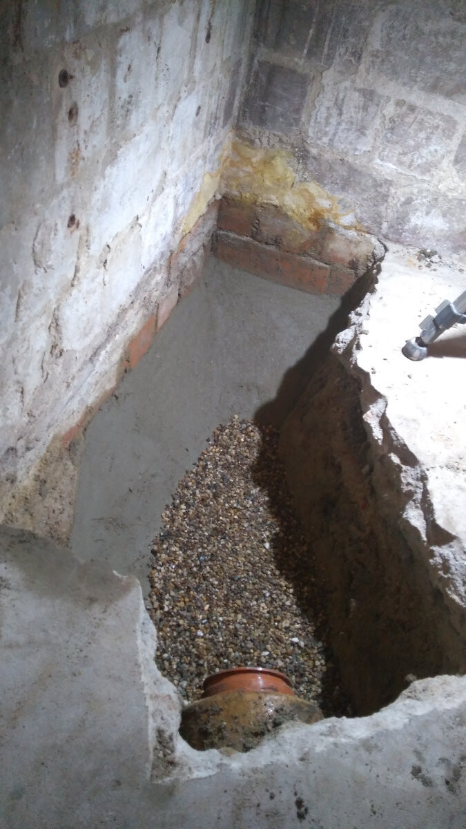 Grube im Betonboden eines Kellers mit Kiesbett, rotem Rohrbogen und bröckelnden Wänden.
