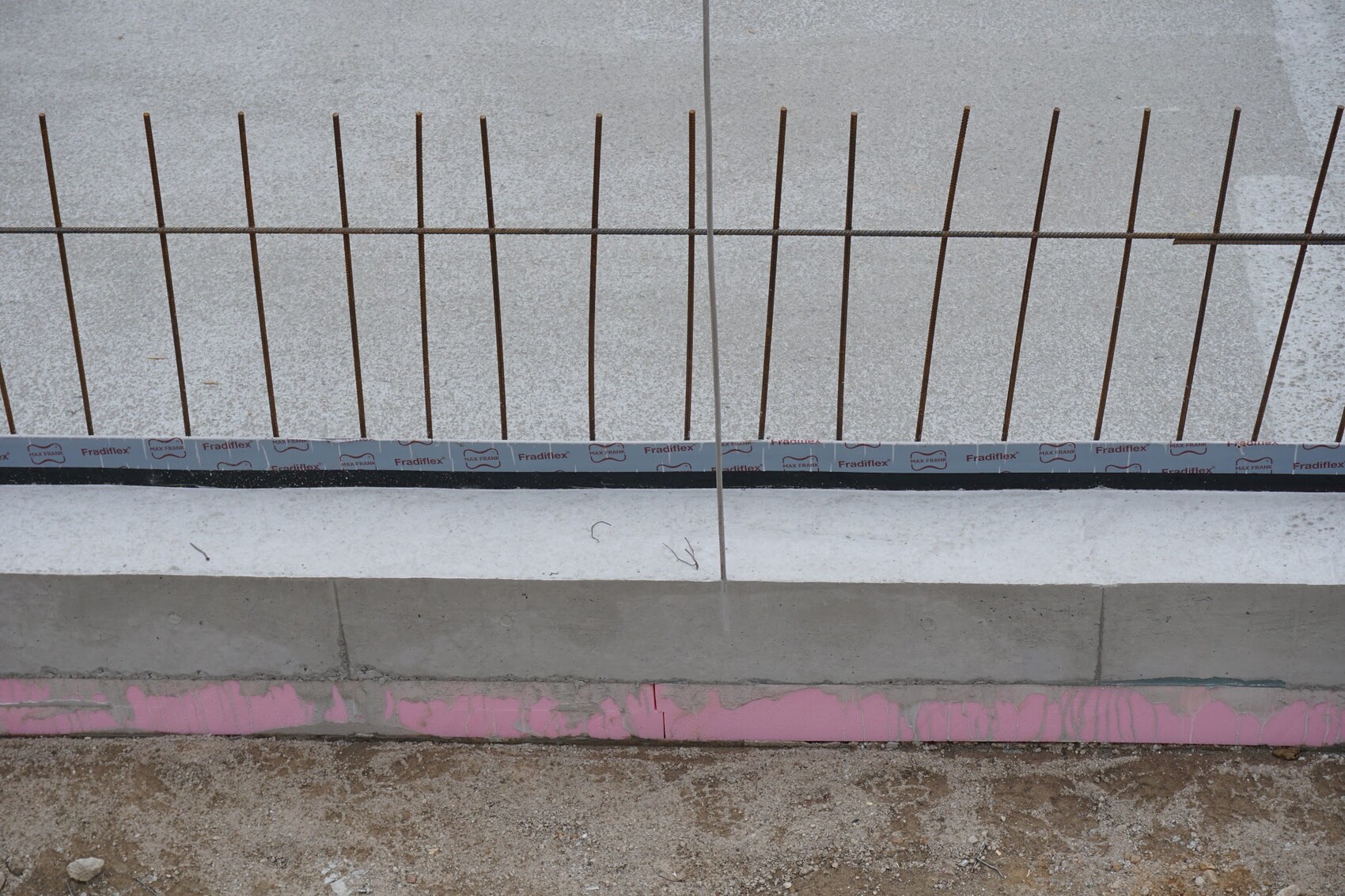 Bewehrungsstahlstäbe ragen aus einer Betonwand; unten rosa Streifen und Baufolie.