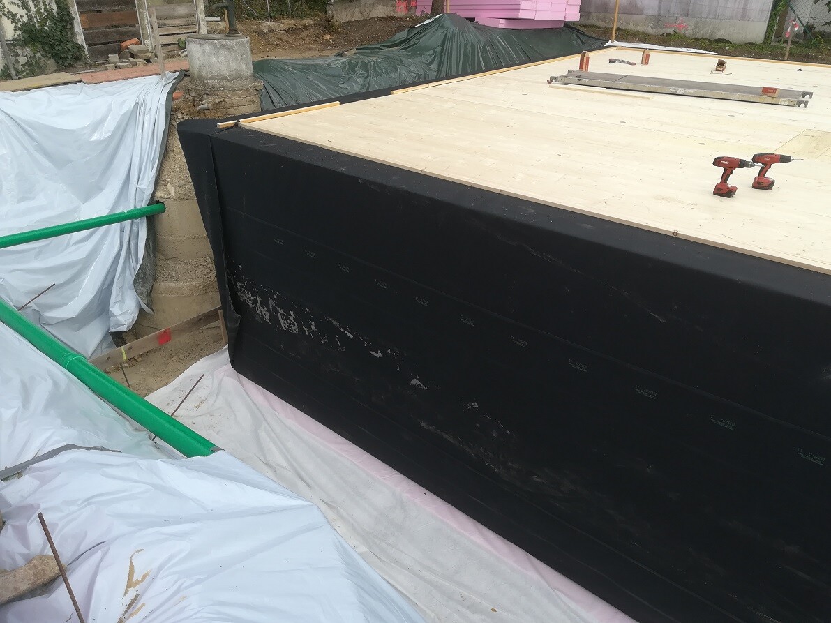 Baustelle mit schwarzer Dichtbahn am Fundament, Holzdeck oben, Werkzeuge auf der Platte.