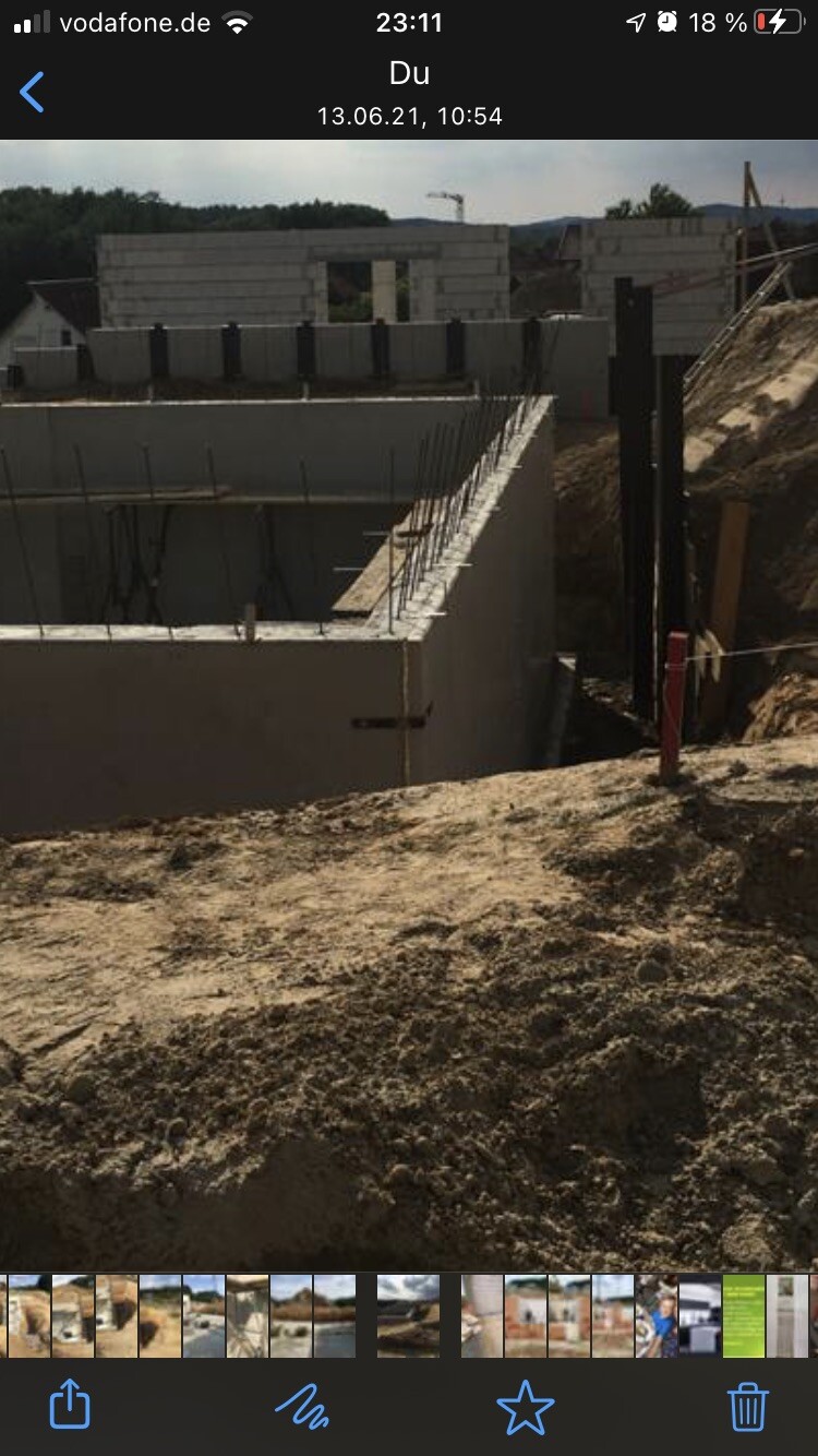Baugrube mit Betonfundament und Stahlbewehrung, Blockmauern im Hintergrund, Erdreich rundherum.