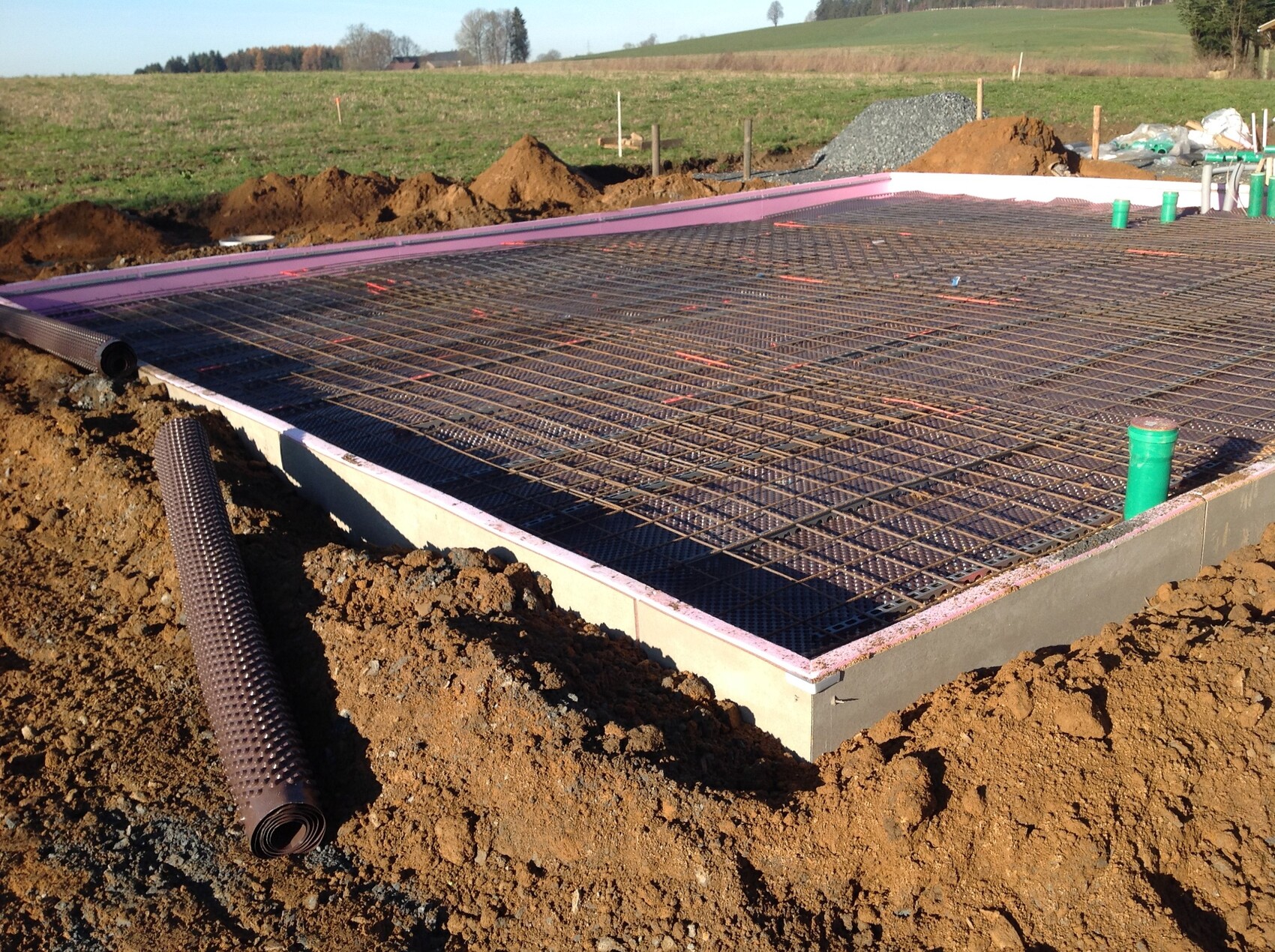 Fundament mit Bewehrungsstahlgitter, Baugrube und grüne Rohre auf Baustelle.