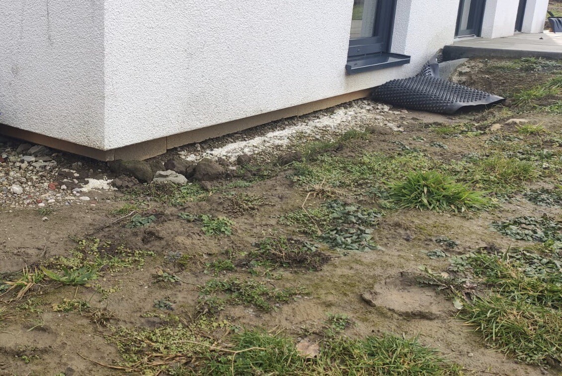 Außenbereich eines Hauses: Fundament unter Putz, Kiesboden, Unkraut und schwarze Drainagematte.
