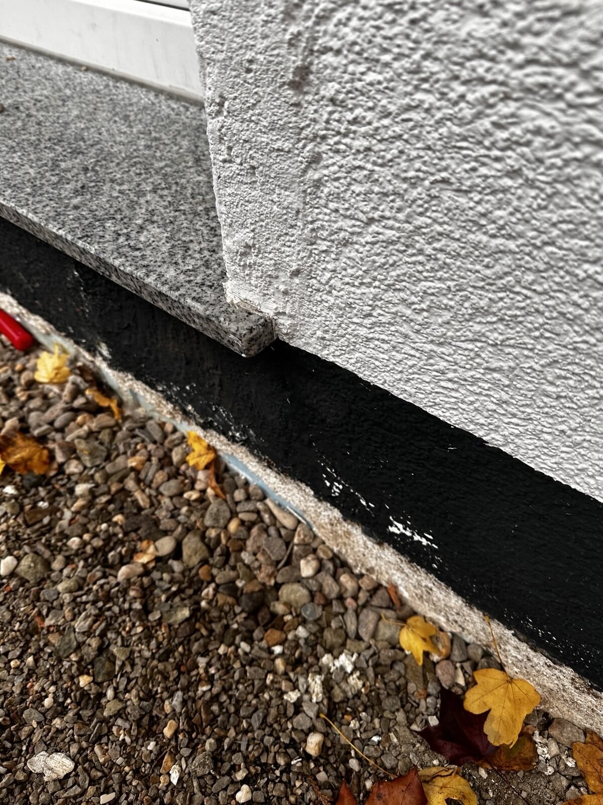 Nahaufnahme einer Fundamentkante mit schwarzer Dichtbeschichtung an der Gebäudewand und Kiesboden.
