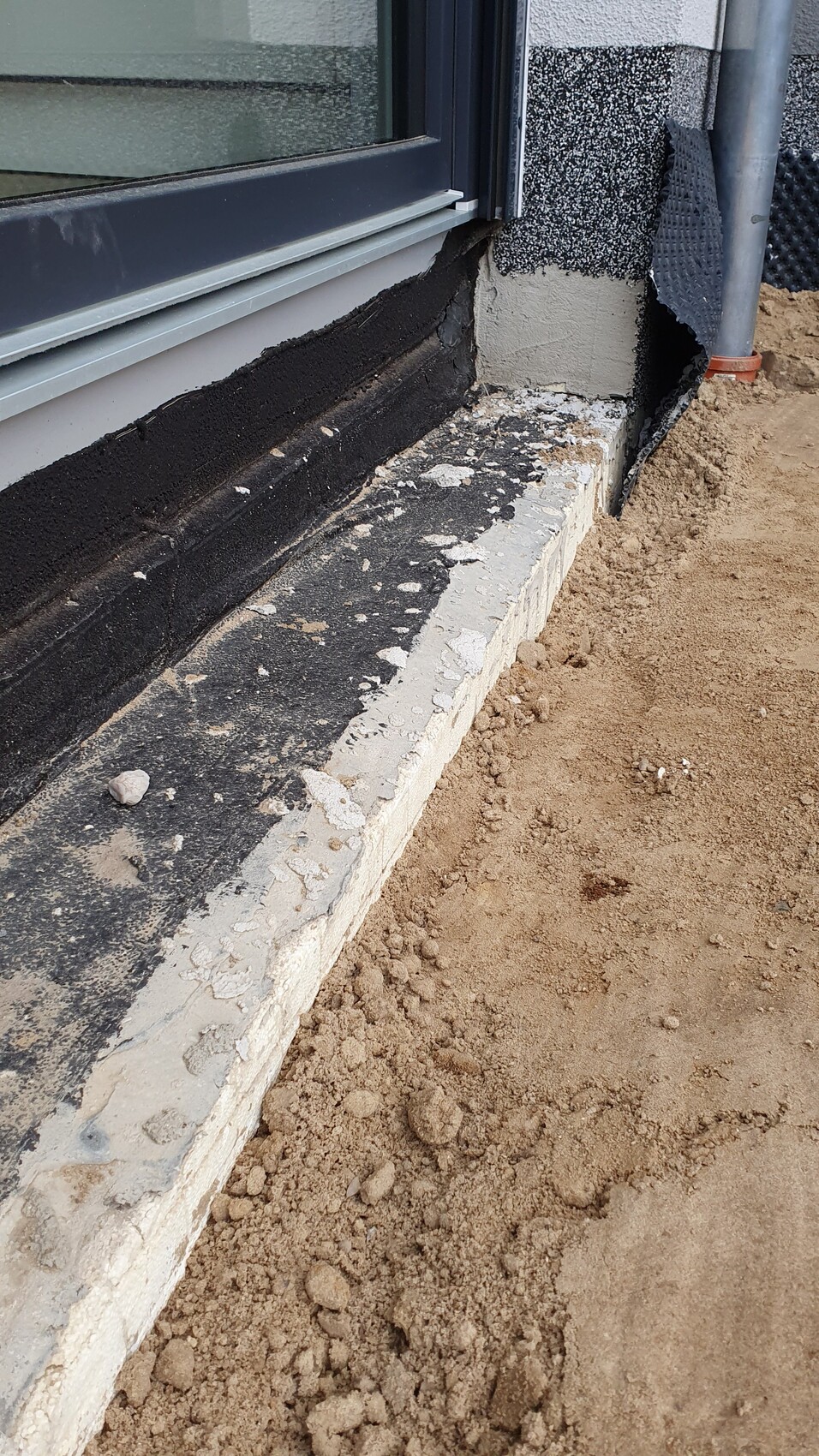 Außenfenster mit schwarzer Abdichtung am Fundament, daneben lose Erdreich und Baureste.