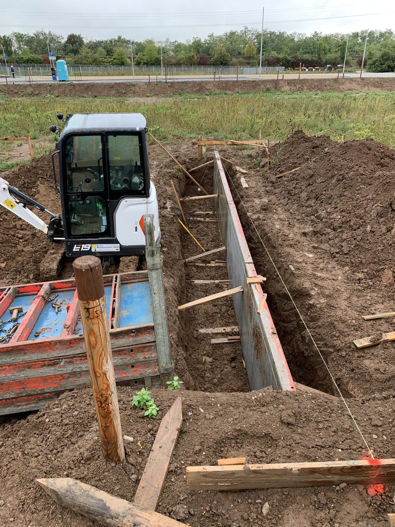 Mini-Bagger in Baugrube neben Betonwand, Holzstützen sichern den Graben.