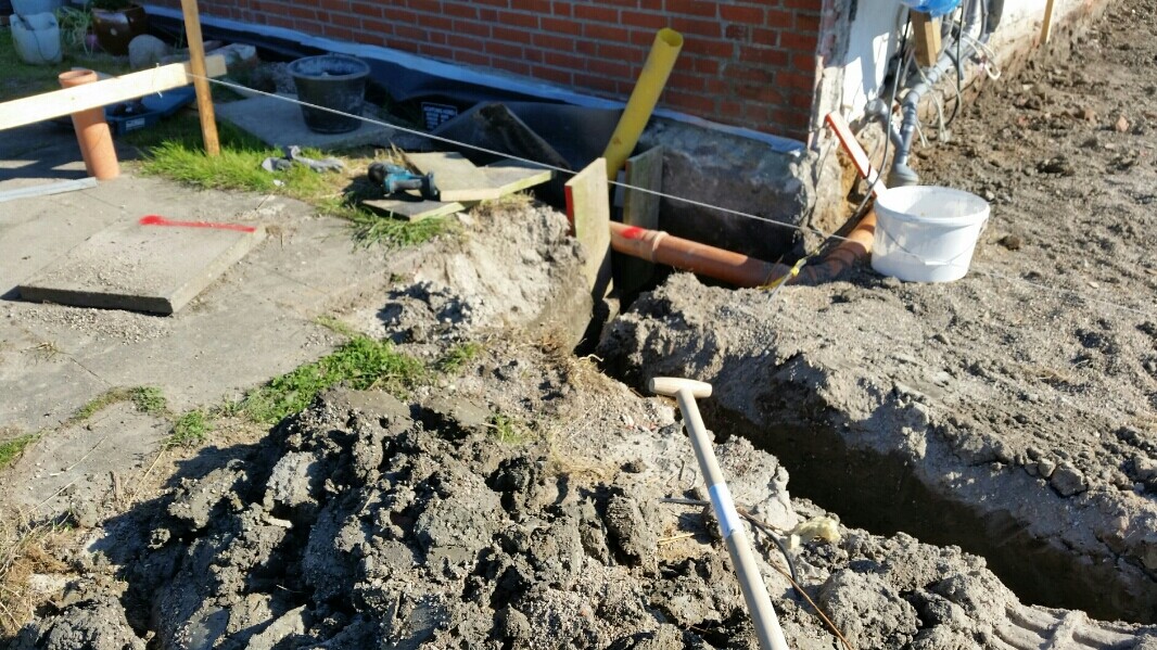 Baustelle mit Graben, Rohrleitungen, Schaufel und Eimer neben Hausfundament