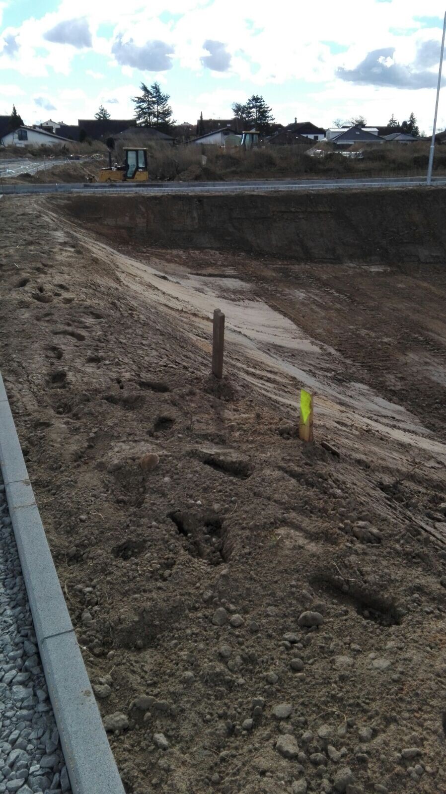 Baustelle mit tiefem Erdgraben, Markierungsstangen und Baufahrzeug im Hintergrund.