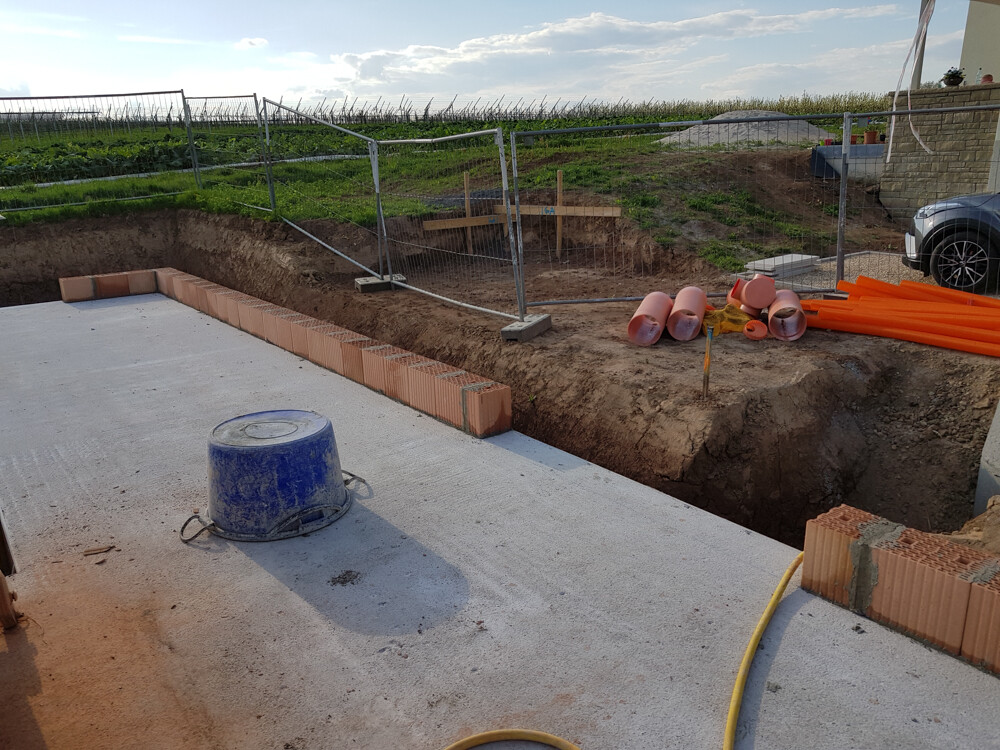 Baustelle: Betonboden, Ziegelrand, orange Rohre, blaue Kanne, Absperrzaun im Hintergrund.