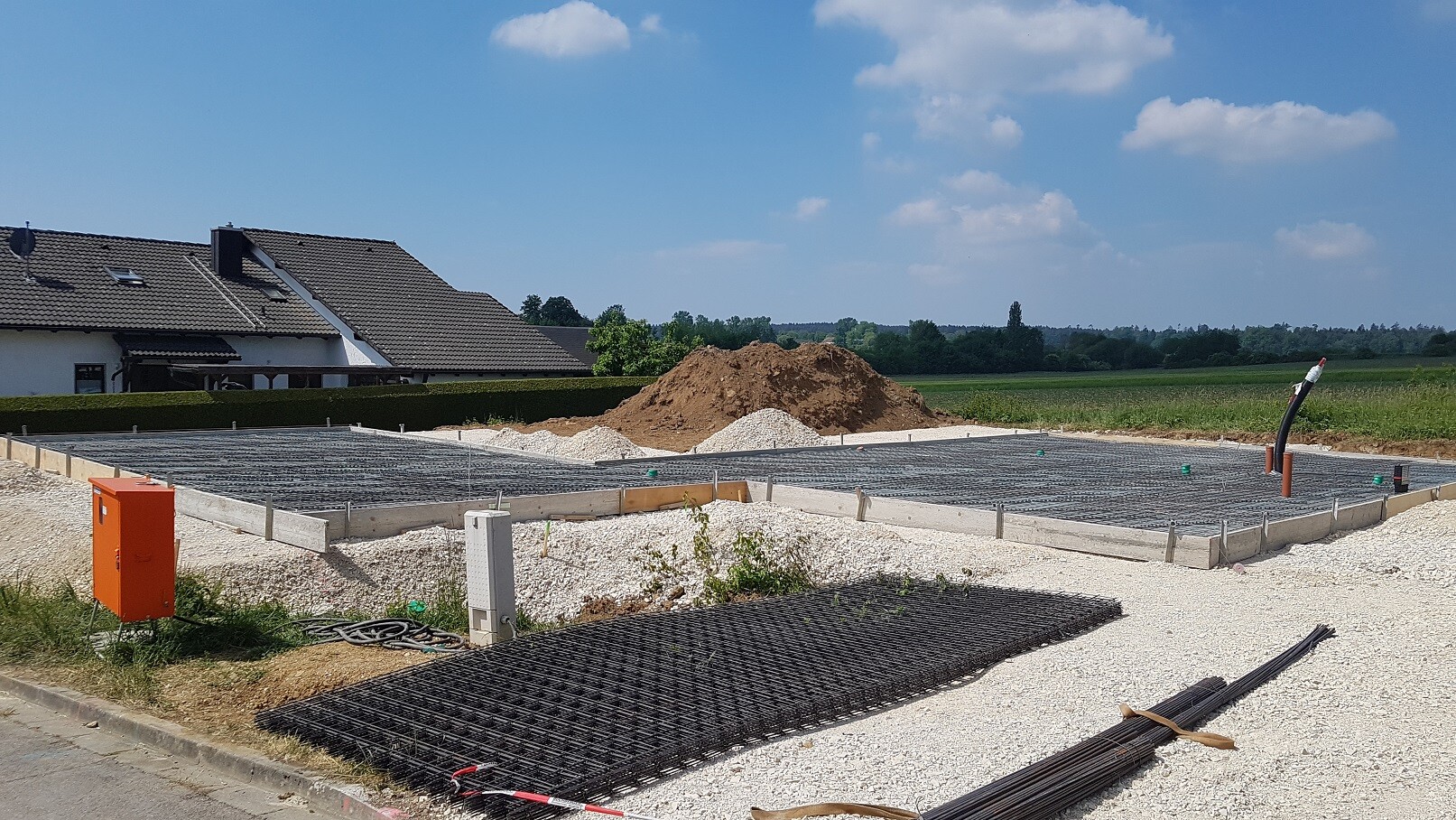 Fundamentbau-Baustelle mit Bewehrungsstahlgitter, Kies und angrenzendem Haus