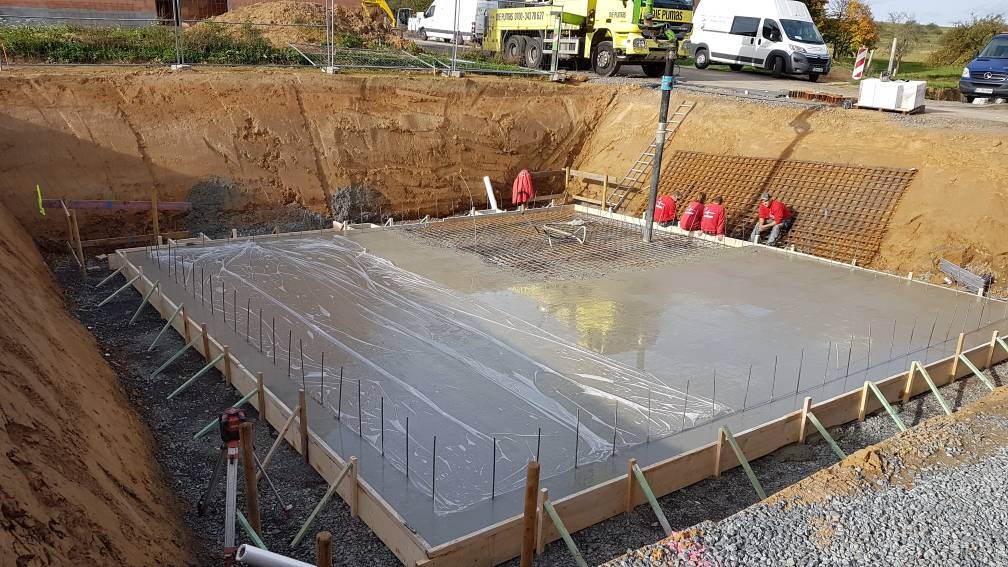 Betonfundament in Baugrube mit Bewehrung, Arbeiter in Rot, Baufahrzeuge im Hintergrund.
