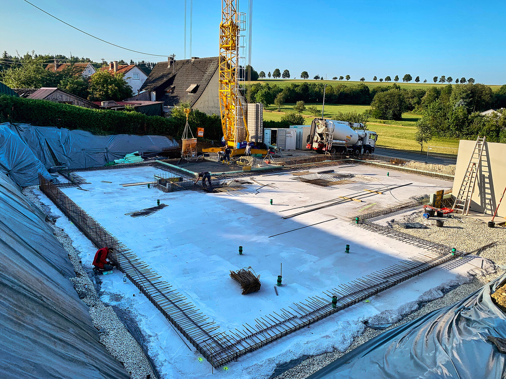 Baustelle: Betonfundament mit Bewehrung, gelber Kran, Betonmischer, Arbeiter.