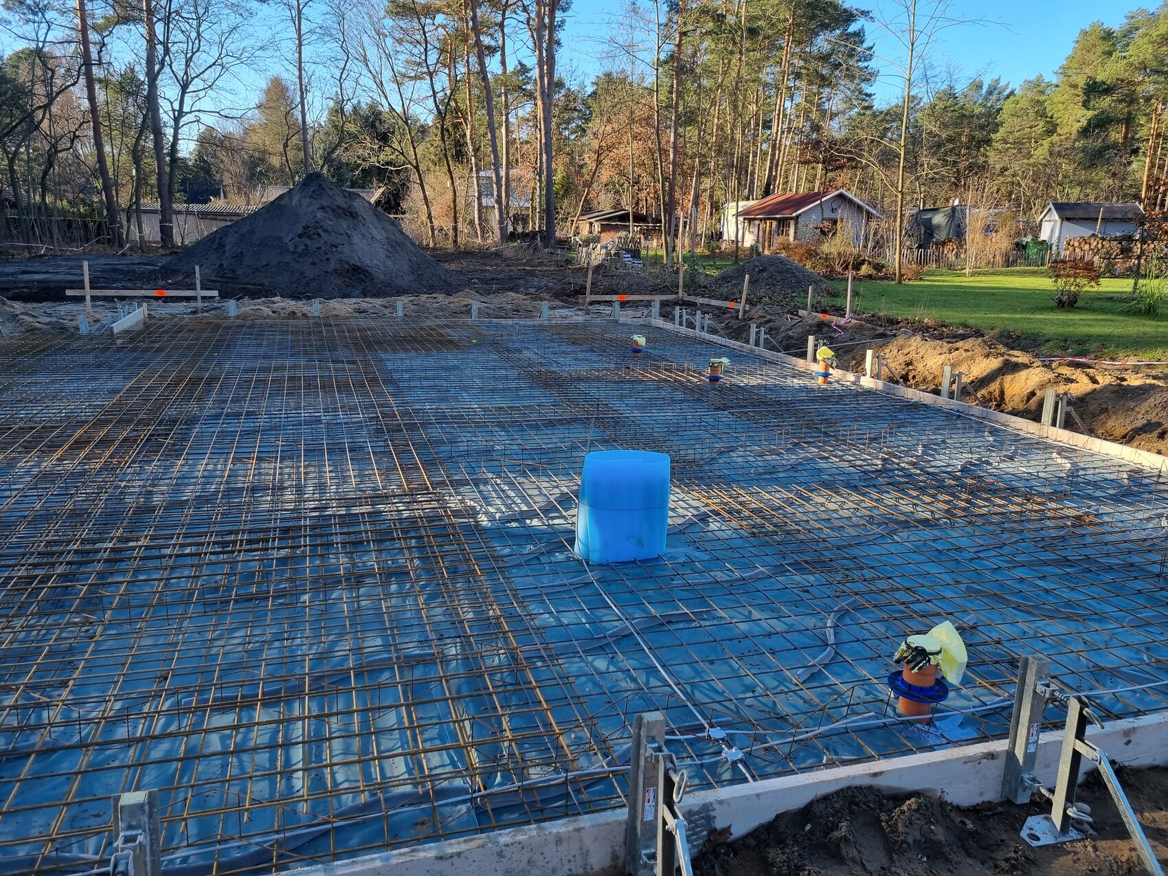 Fundament mit Stahlbewehrung auf Abdeckfolie; blaue Zisterne, Baugrube, Wald im Hintergrund.