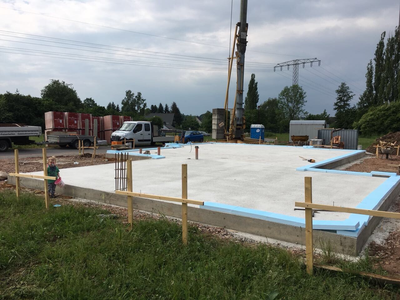Fundamentplatte auf Baustelle mit blauer Dämmung, Holzpfosten und Kleinkind am Rand.