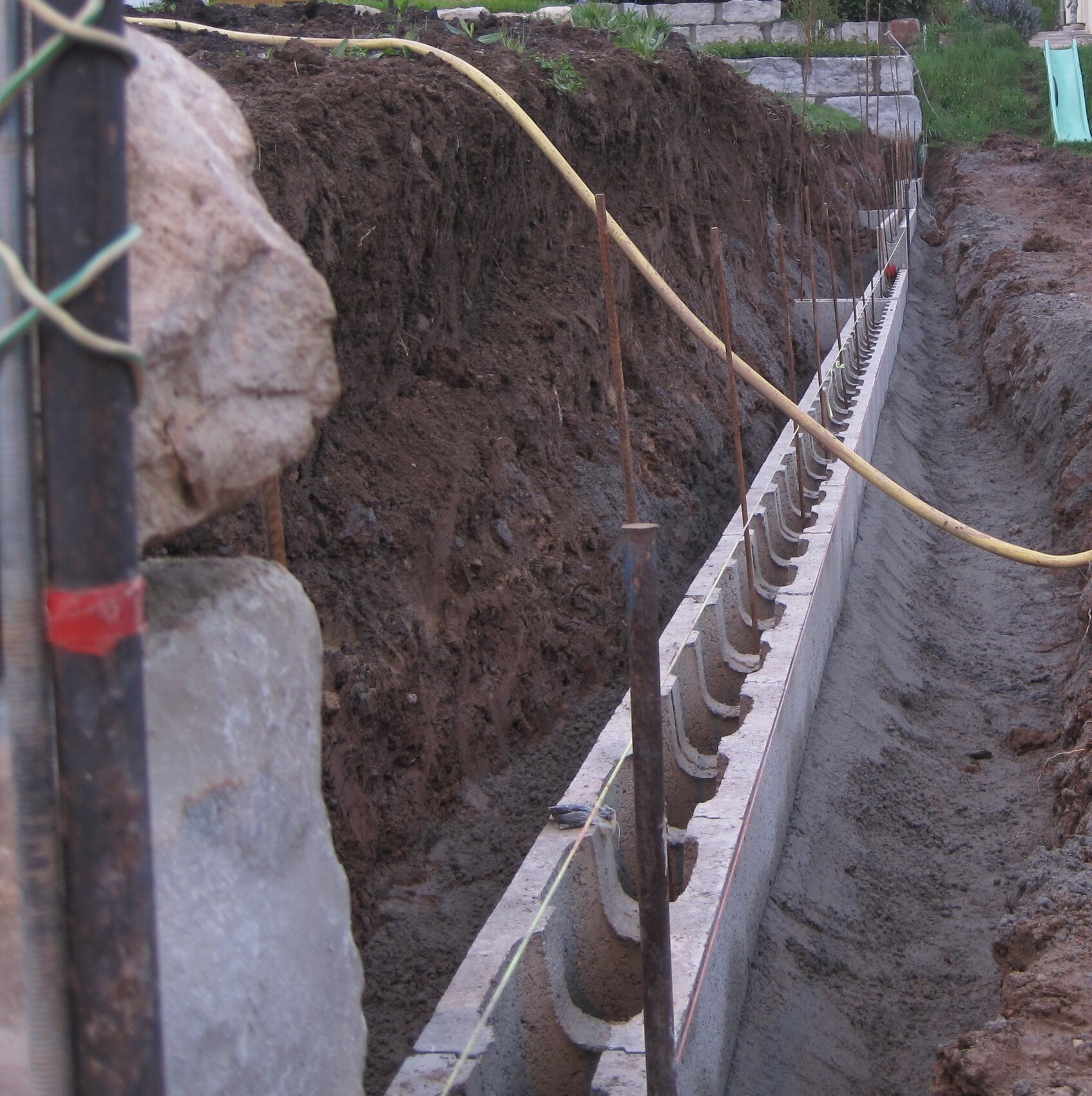 Graben für Fundament mit Betonwand, Bewehrungstangen und Baugeräten sichtbar