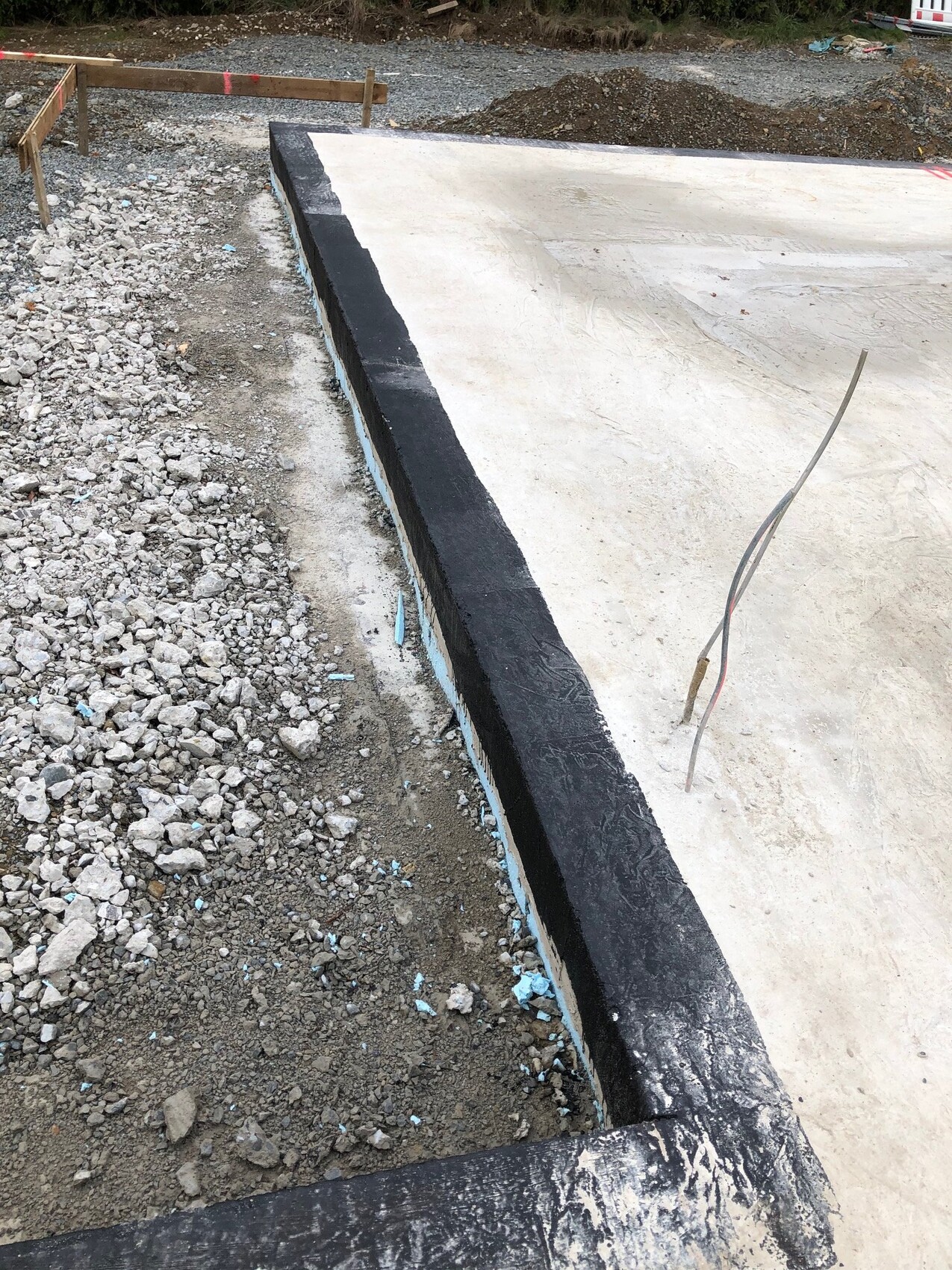 Betonfundament mit schwarzer Abdichtung am Rand, umliegender Kies und Baubaustelle.