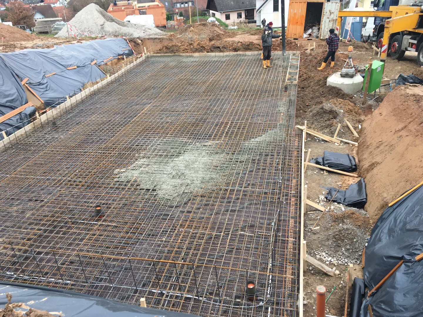 Baustelle mit Bewehrungsstahlnetz für Betonboden; Arbeiter in Gelbstiefeln, Geräte im Hintergrund