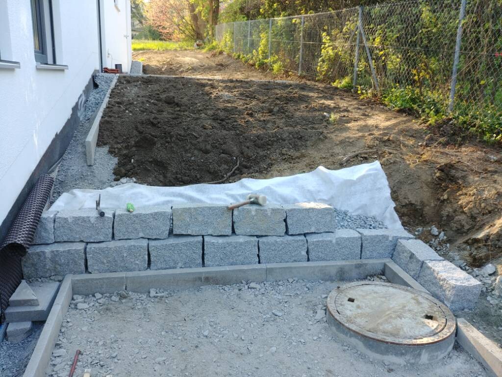 Stützmauer aus Betonwürfeln neben einem Haus, Baugrube dahinter, runder Schachtdeckel sichtbar.