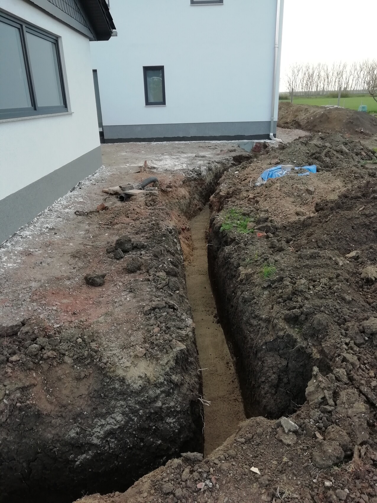 Baugraben neben weißem Haus; Erdarbeiten, Sandboden sichtbar, Rohre liegen bereit.