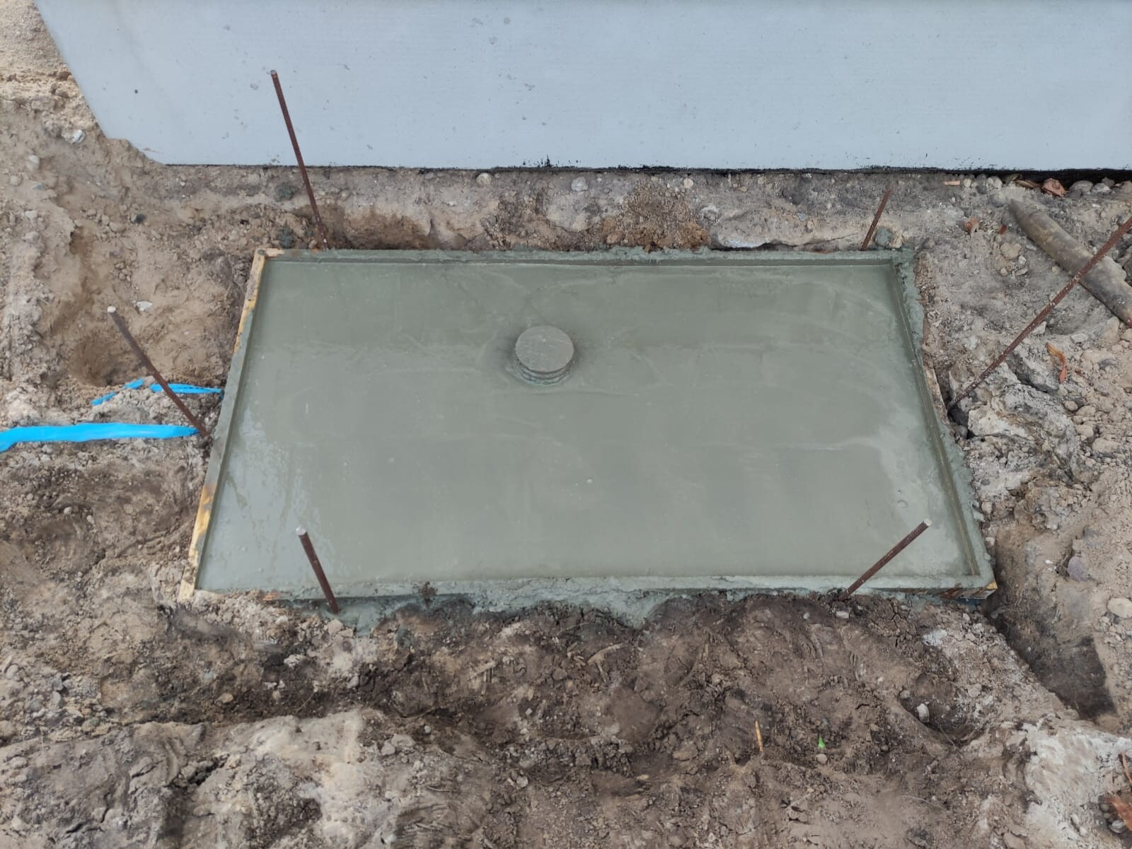 Frisch gegossene Betonplatte in Grube mit zentralem Rohrdeckel und Bewehrung.