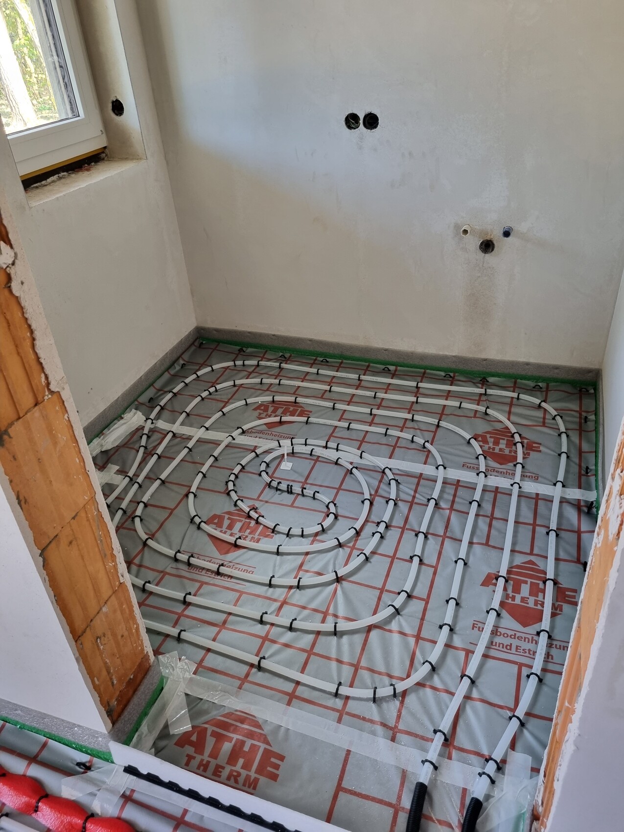Unterflur-Heizung: Spiralförmige Rohre auf Dämmfolie im Bau-Badezimmer, Wandanschlüsse sichtbar.