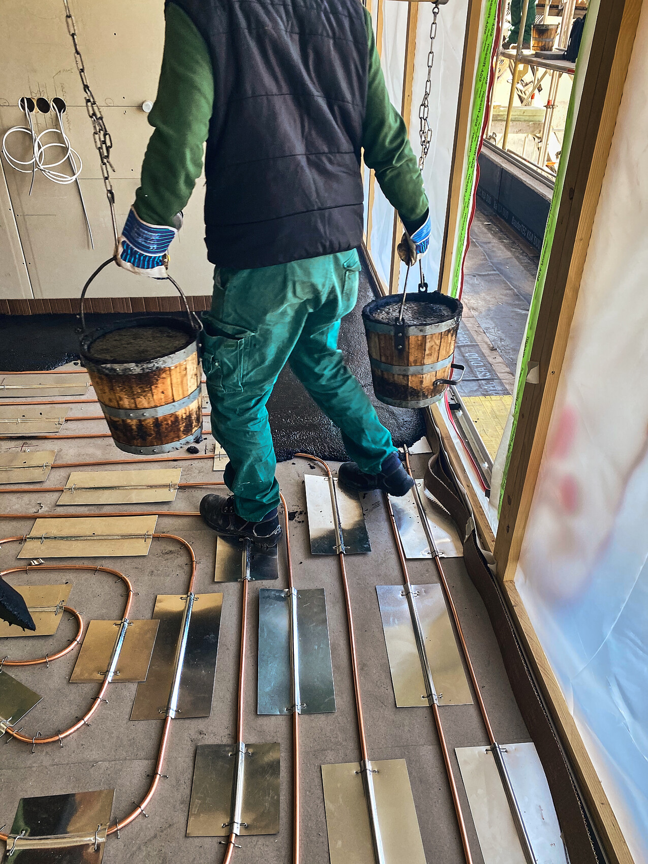 Bauarbeiter trägt zwei Eimer Mörtel; Boden mit Kupferrohren und Metallplatten.
