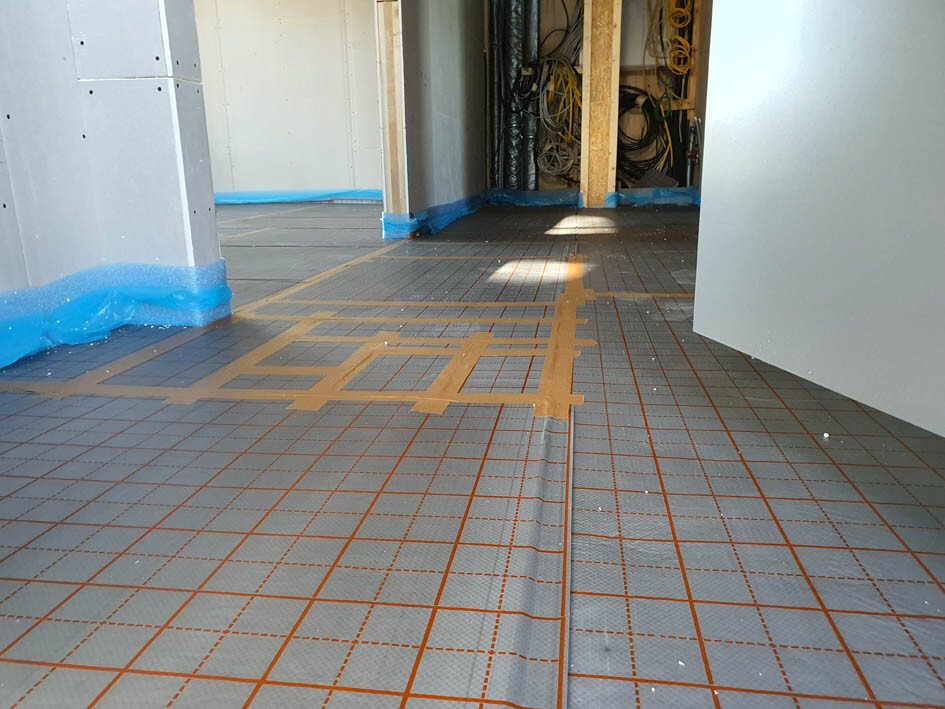 Baustelle: Verlegte Bodenheizung-Matten, orange Linien, blaue Schutzfolie, unfertiger Innenraum.