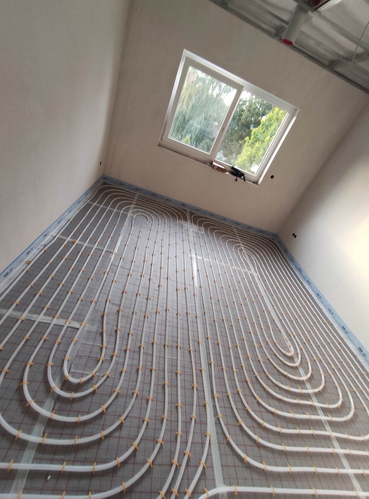 Raum mit Fußbodenheizung: weiße Rohre in Spiralen auf dem Estrich, Fenster sichtbar.