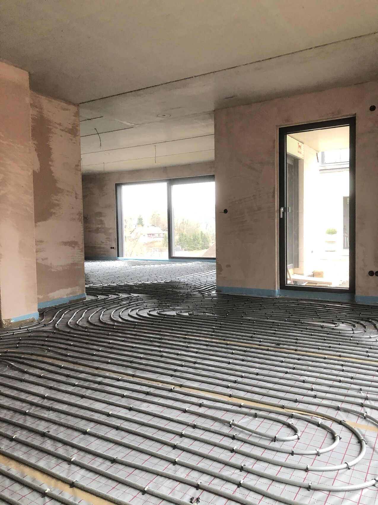 Innenraum einer Baustelle: verlegte Fußbodenheizungsrohre, Fensterfront und Tür nach draußen.