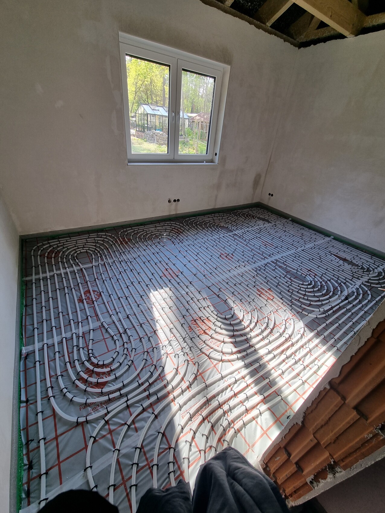 Rohbauzimmer mit verlegter Fußbodenheizung: Heizrohre in Spiralen auf Unterlage, Fenster sichtbar.
