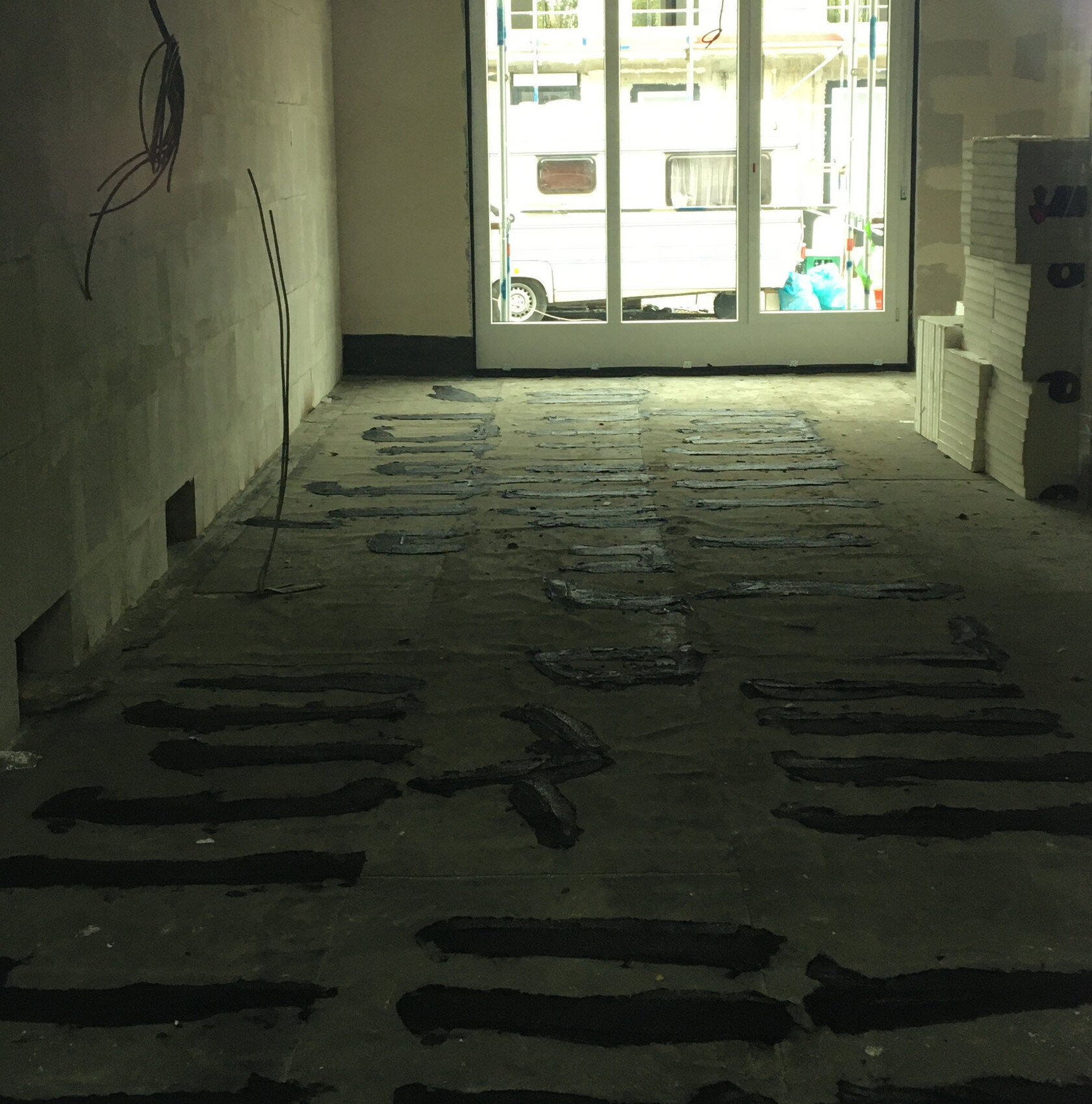 Innenraum einer Baustelle: roher Boden mit schwarzen Abdichtungsstreifen, Fensterfront nach draußen.