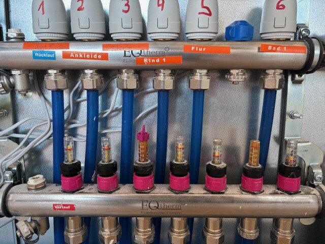 Verteiler für Heizkreise: sechs blaue Rohre, pinke Verschraubungen, Nummern 1–6.