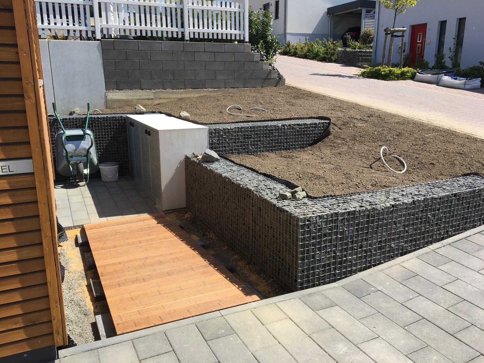 Gartenbaustelle mit Gabionen-Wand, Erdarbeiten, Holzrampe und Schubkarre.