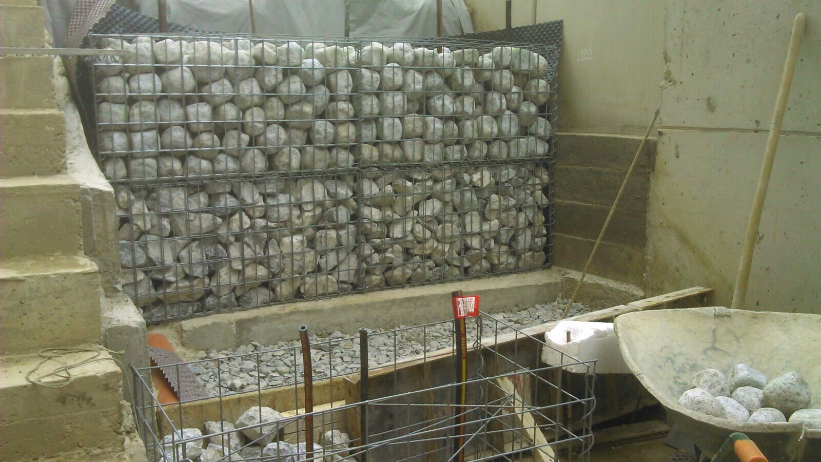 Gabionenwand aus Drahtkörben mit Steinen; Baustelle mit Schubkarren und Betonarbeiten.