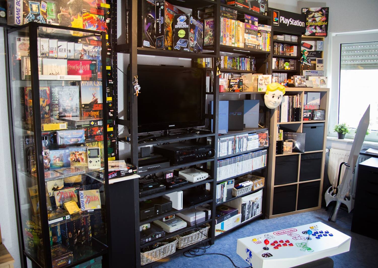 Retro-Gaming-Zimmer mit Regalen voller Konsolen, Box-Spielen, TV und Vault Boy-Maske.