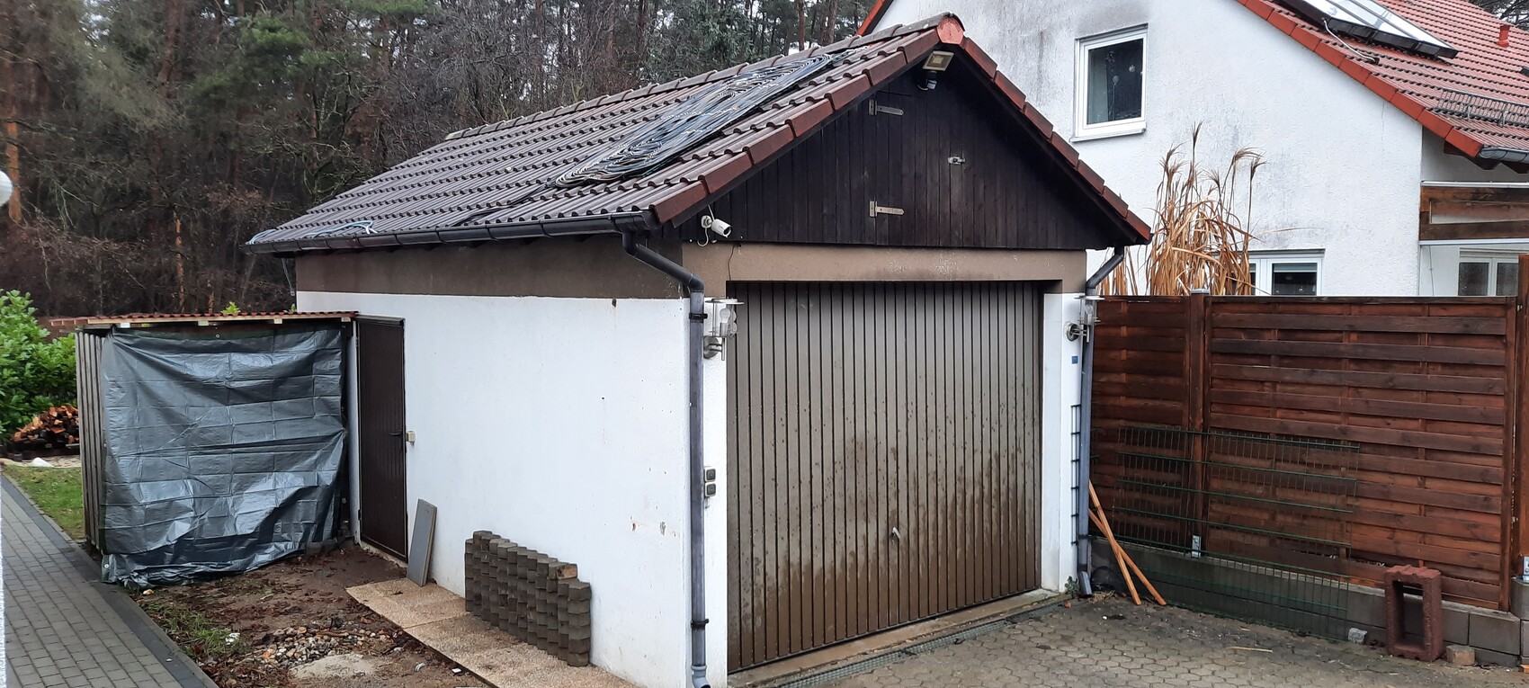 Kleiner Garagenanbau mit braunem Holztor, linkem Schuppen mit grauer Plane, Holzlatten-Zaun rechts.