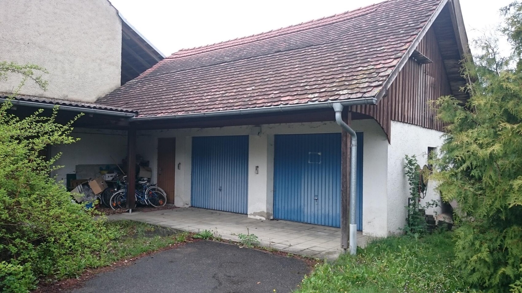 Außenansicht eines Gebäudes mit zwei blauen Garagentoren, Fahrradabstellbereich und Dachüberstand.
