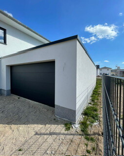 Moderne weisse Garage mit dunkelgrauem Garagentor, Einfahrt aus Kies, Zaun rechts.
