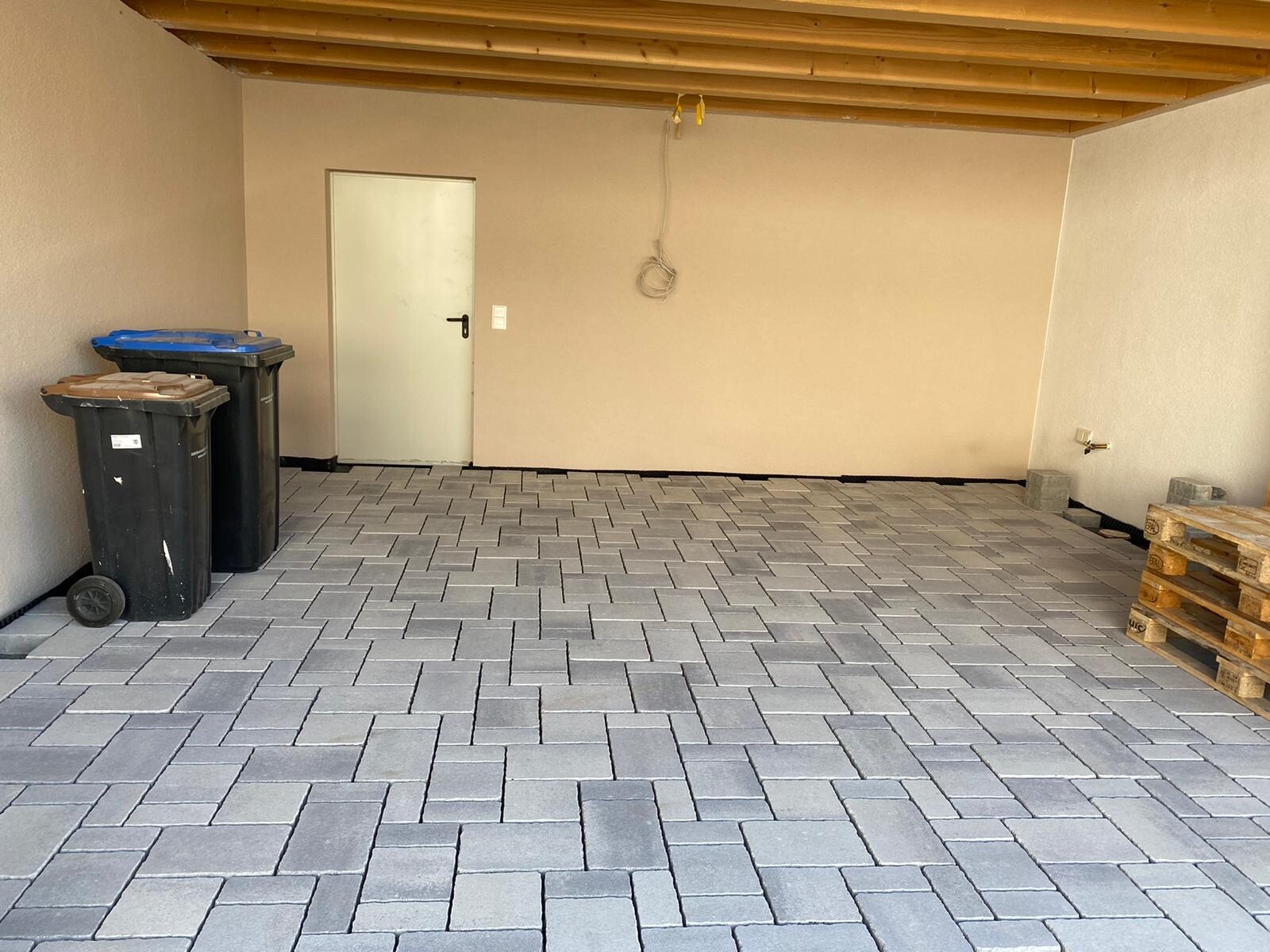 Gepflegter Garage mit Pflasterboden, Mülltonnen links, Paletten rechts, Tür hinten.