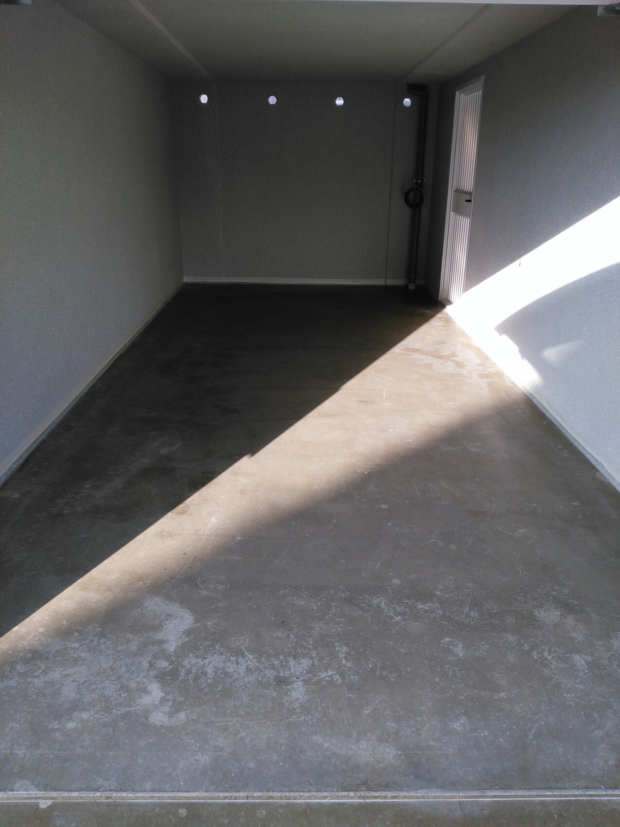 Leerer grauer Abstellraum/Garage mit Betonboden, Tür rechts, diagonales Sonnenlicht.