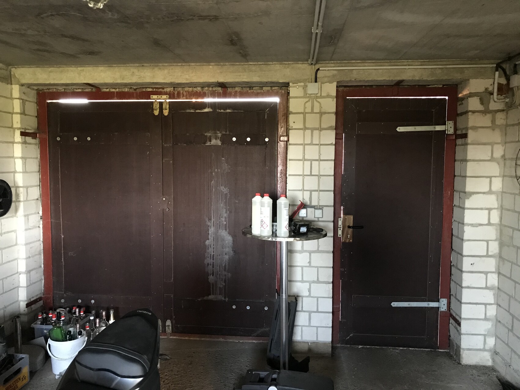 Garage/Schuppen mit dunklen Holztüren, zentraler Tisch mit Reinigungsflaschen, Motorradbank links.