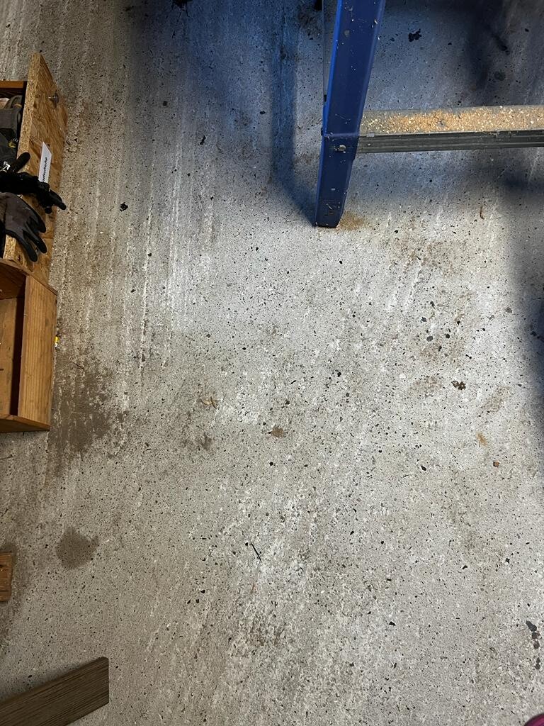 Betonboden in Werkstatt mit Flecken; linke Holzkisten mit Handschuhen, rechte blaue Leiter.