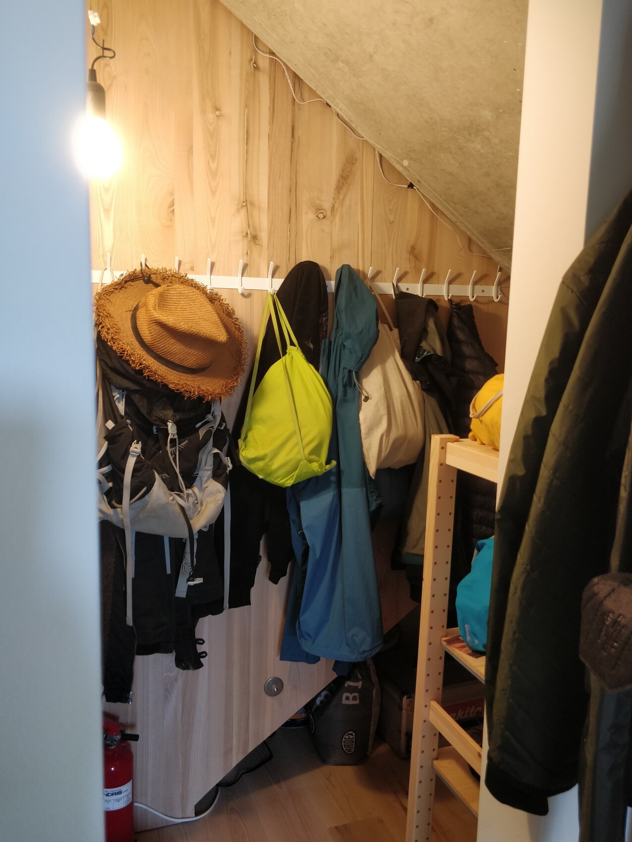 Kleiner Garderobenraum mit Hakenleiste, Jacken, neon-grüner Rucksack, Hut, Glühbirne.
