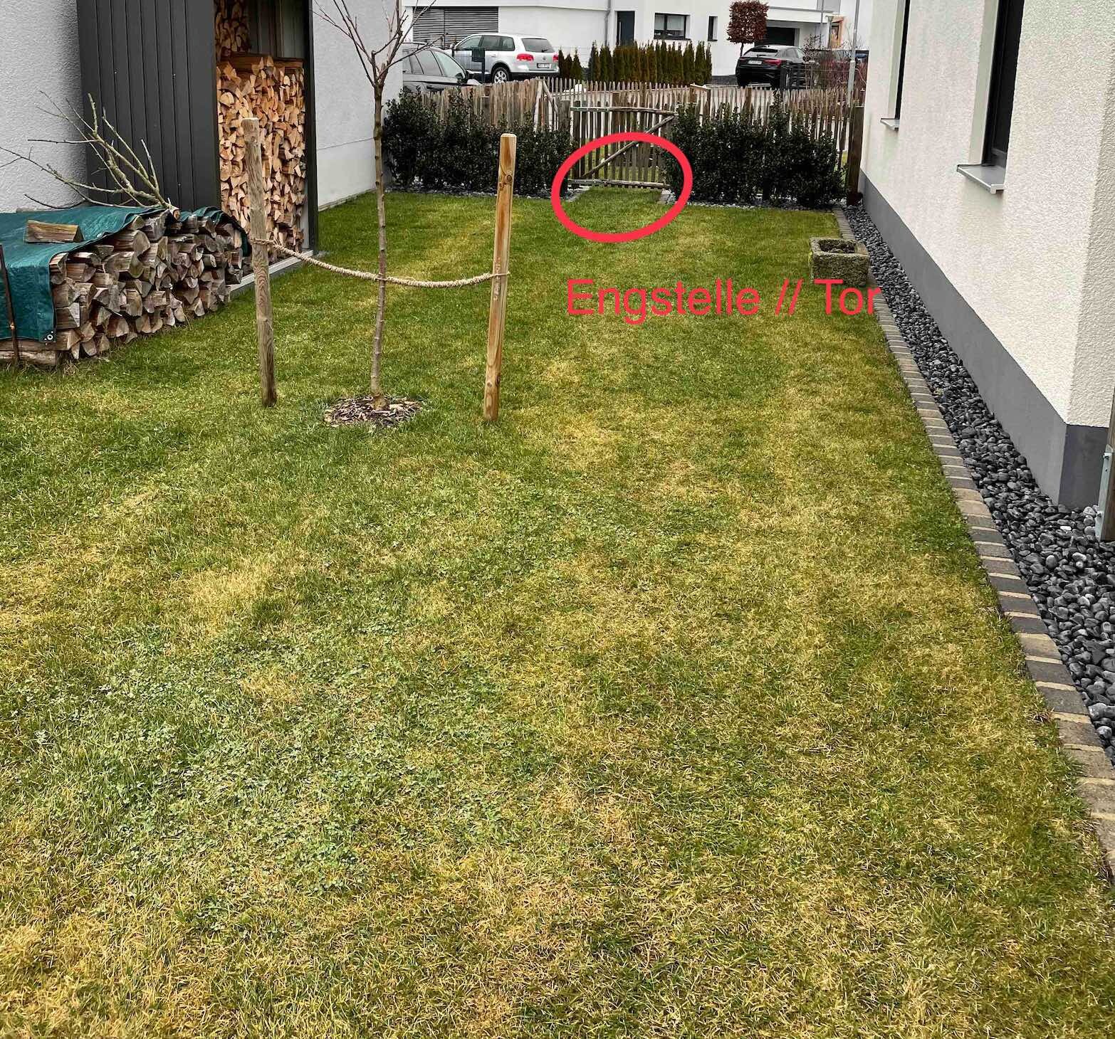 Garten mit Gras, zwei Holzpfosten und Seil als Begrenzung, Brennholz links, Zaun hinten.