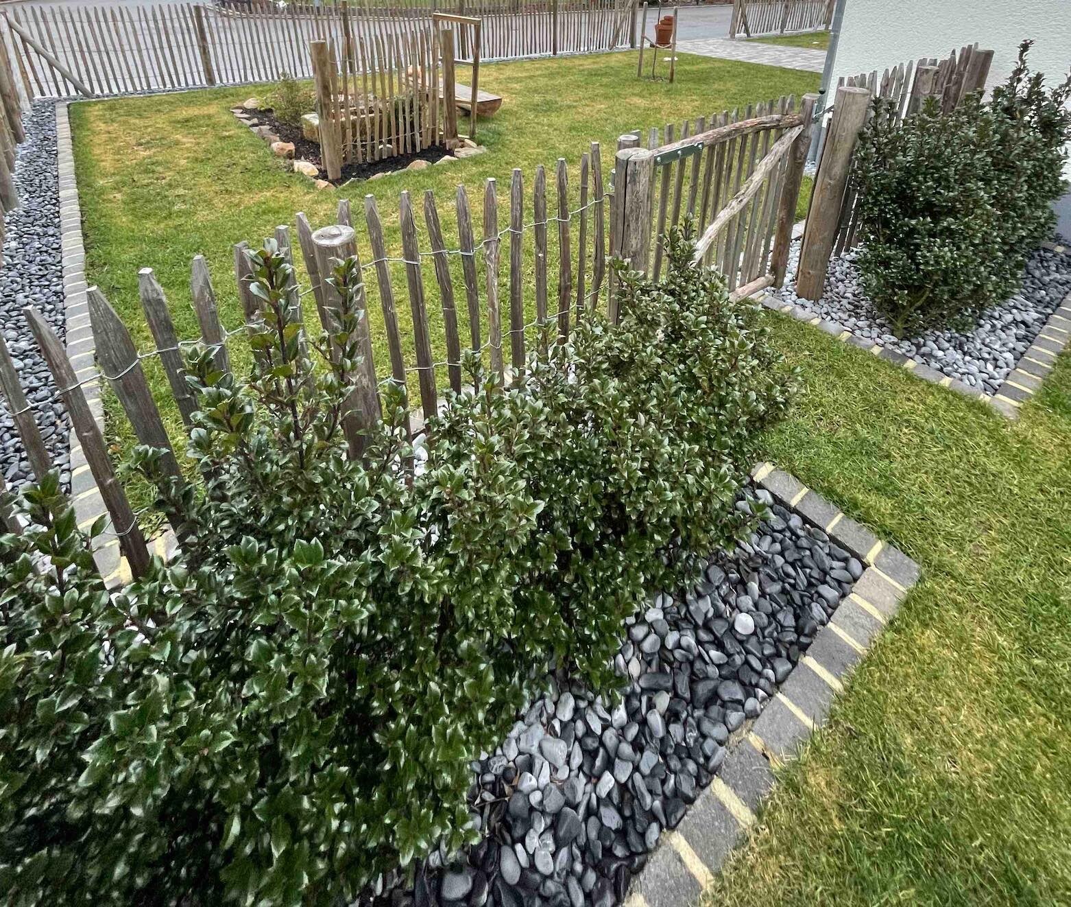 Garten mit Holzpalisadenzaun, dichter grüner Hecke, Kieselboden und Grasfläche