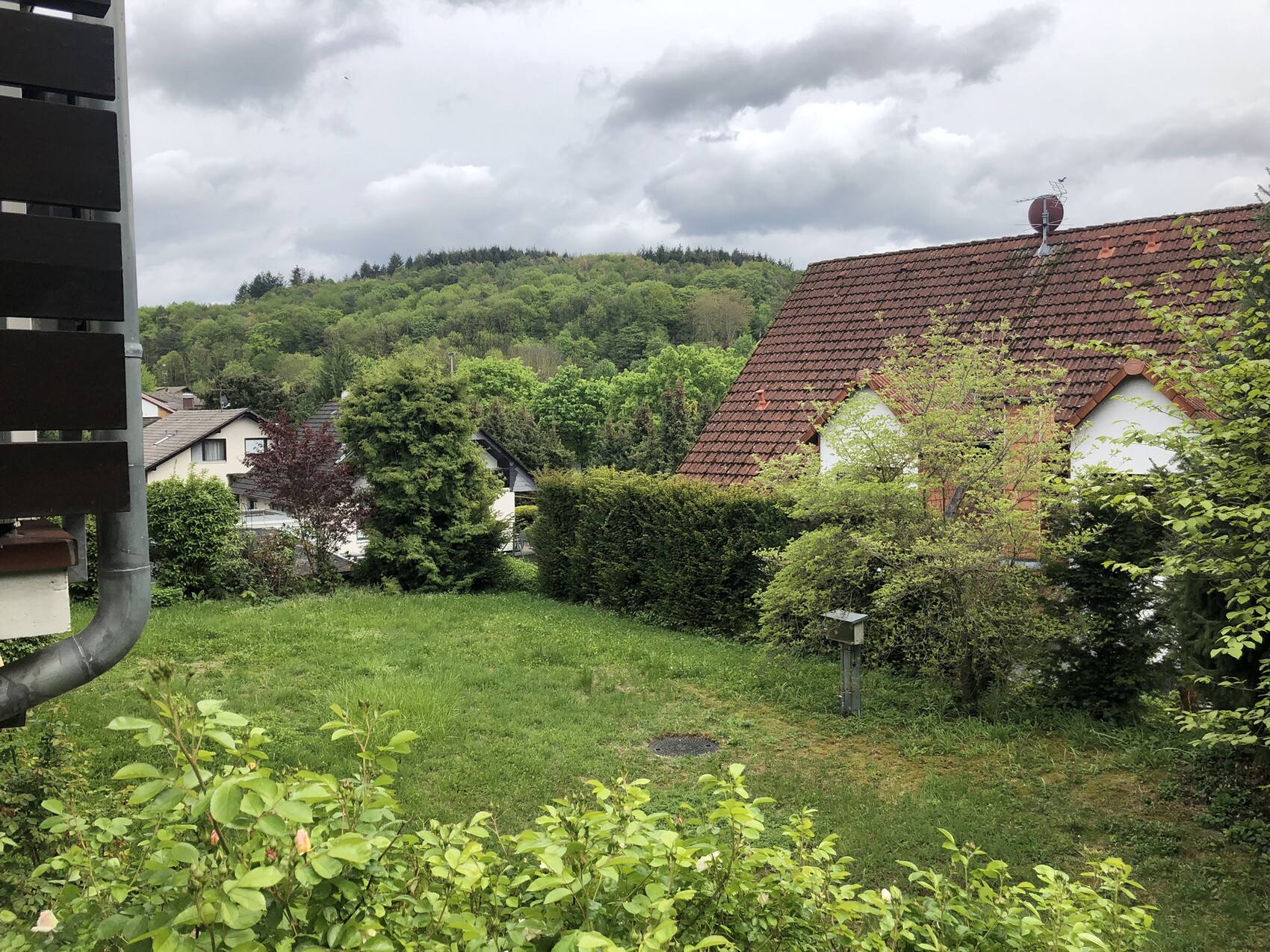Garten mit Gras, Sträuchern; Haus mit rotem Ziegeldach, bewaldeter Hügel im Hintergrund.