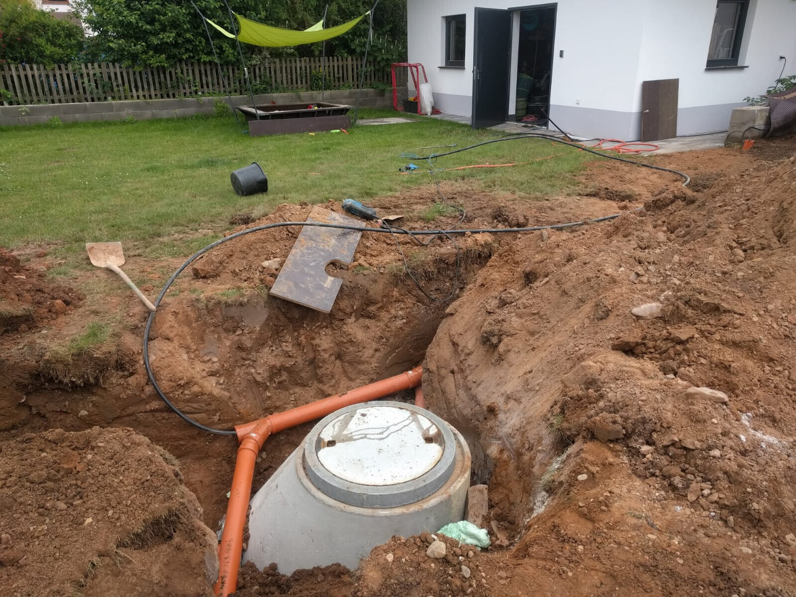 Graben im Garten mit Revisionsschacht aus Beton orange Abwasserrohre Werkzeuge Haus im Hintergrund
