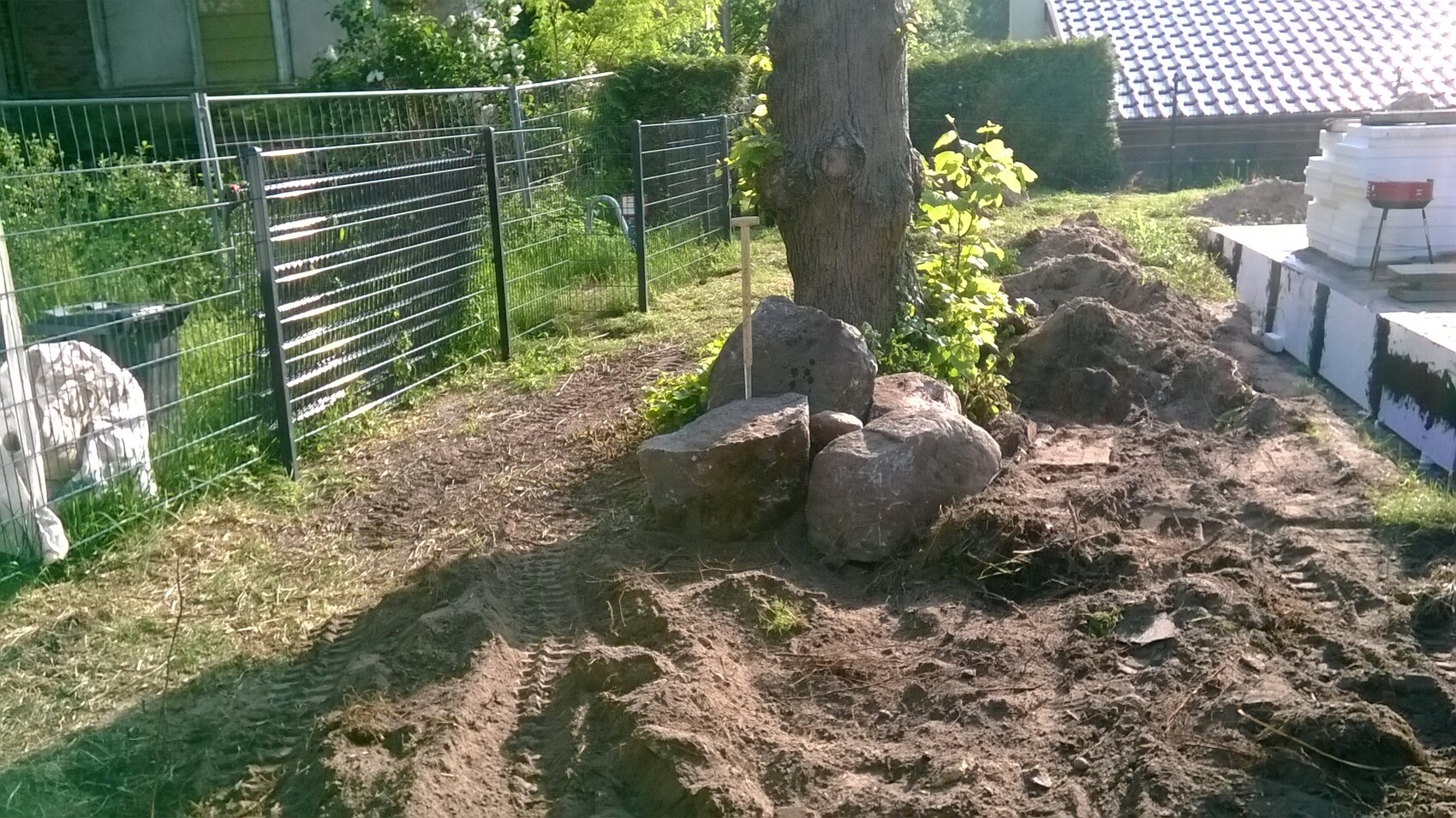 Baum im Garten mit großen Steinbrocken um den Stamm, aufgeschüttete Erde und Baumaterialien.