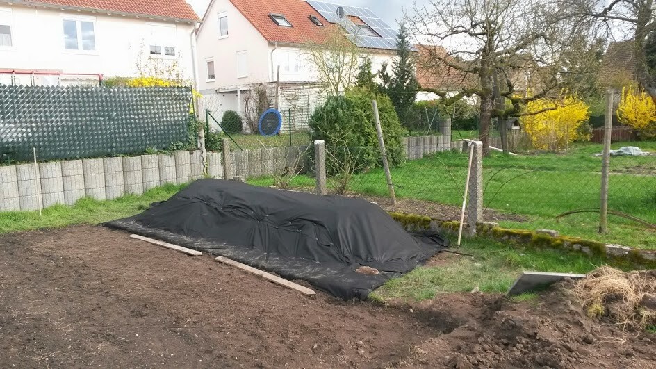 Garten hinter Häusern: schwarze Abdeckplane deckt einen Erdhügel ab, Zaun, Sträucher und Bauholz liegen daneben.