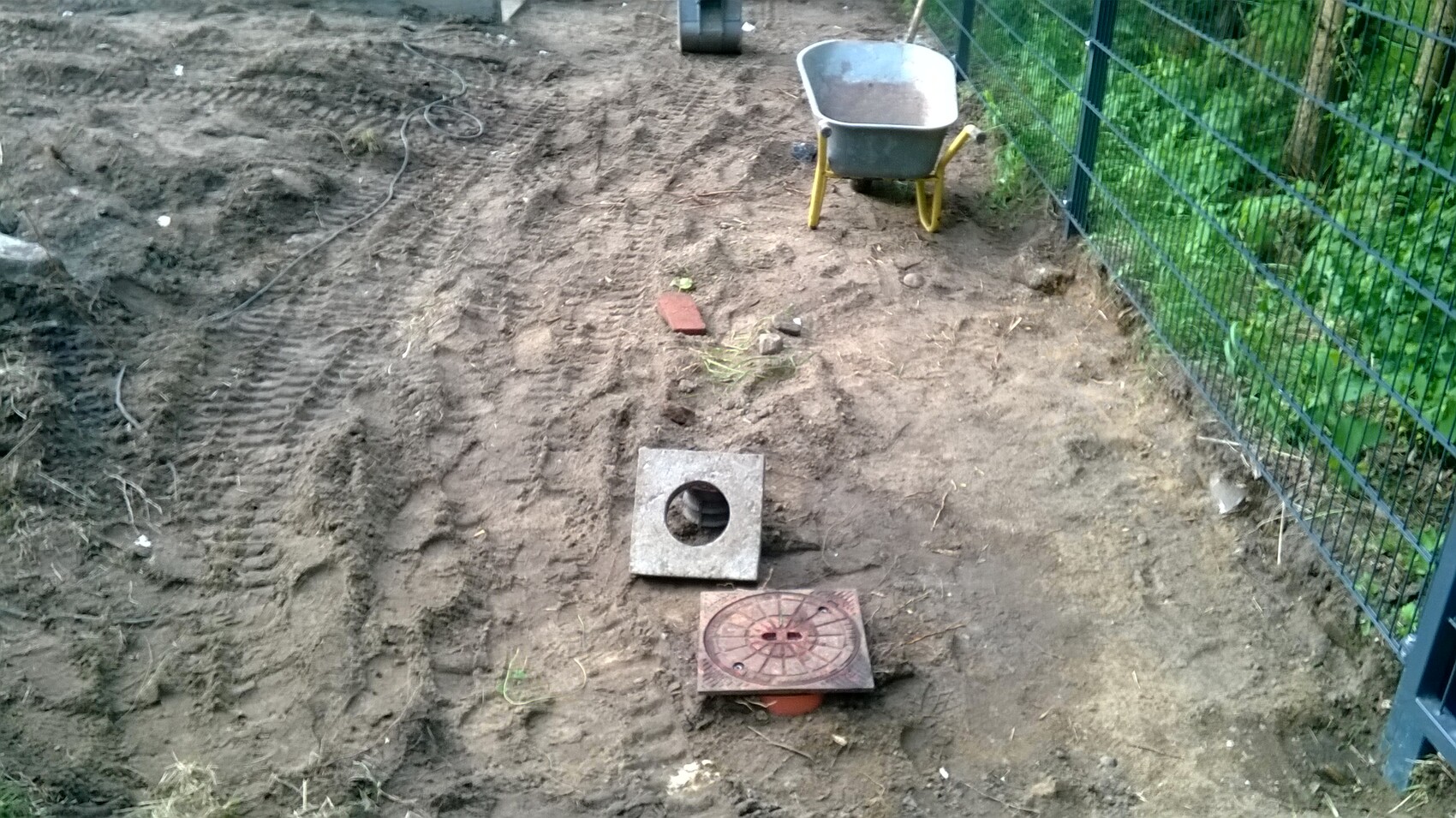 Gartenbaustelle mit freigelegter quadratischer Betonplatte mit Loch, Schubkarre und Zaun.