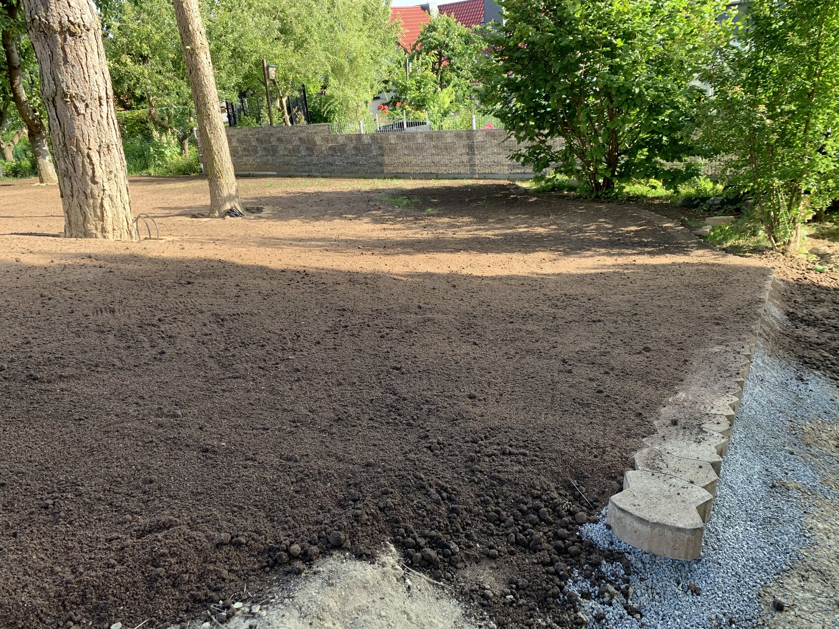 Gartenfläche mit dunkler Erde, links Bäume, rechts Steinmauer im Hintergrund.
